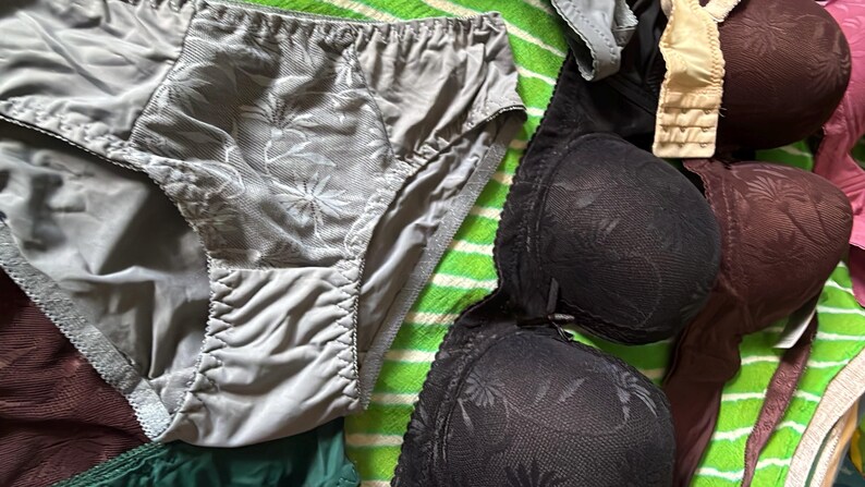 May include: A collection of undergarments including a pair of grey briefs with a floral lace front panel, a black molded cup bra with a floral pattern, and a dark brown molded cup bra with a matching floral texture and beige shoulder straps.