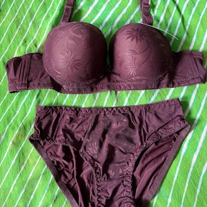 May include: A dark purple lingerie set consisting of a molded cup bra and matching briefs. Both pieces feature a tonal floral pattern on a textured fabric, with adjustable shoulder straps on the bra and a central panel of floral lace on the briefs.