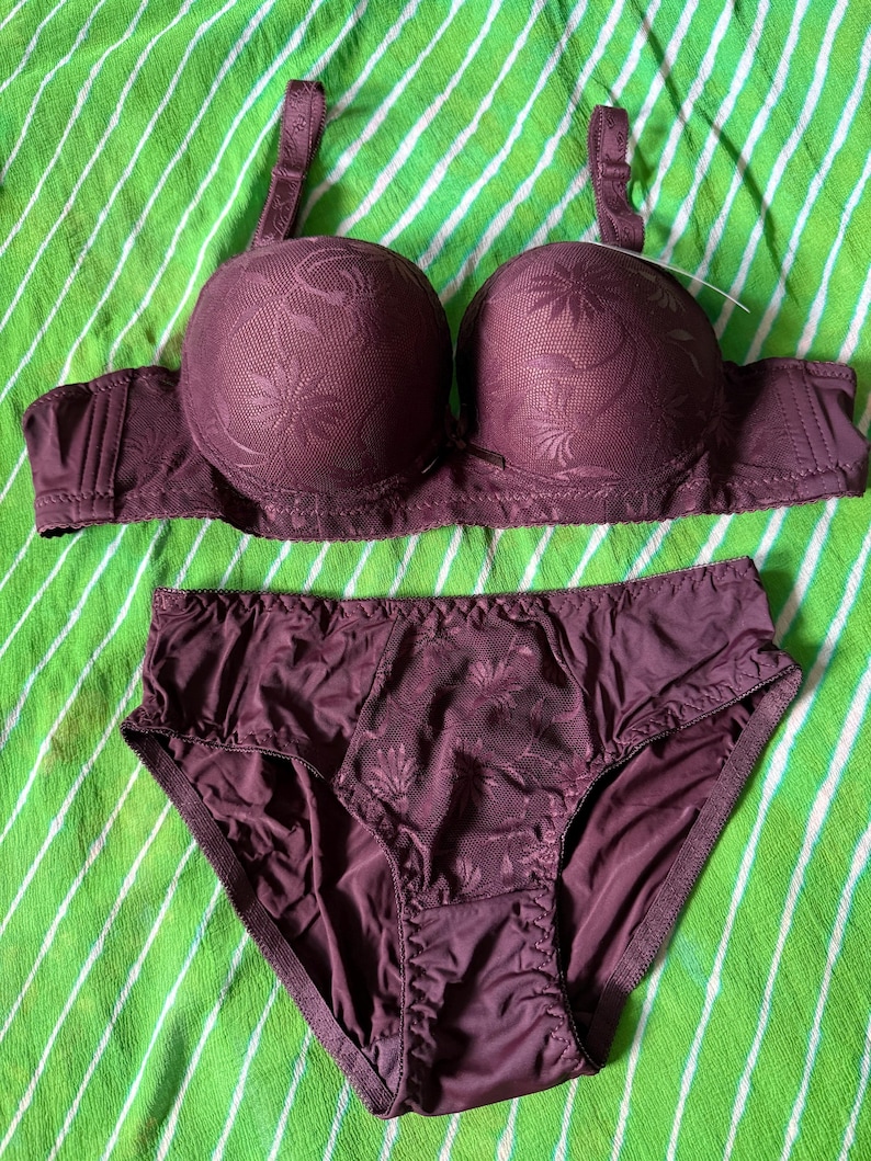 May include: A dark purple lingerie set consisting of a molded cup bra and matching briefs. Both pieces feature a tonal floral pattern on a textured fabric, with adjustable shoulder straps on the bra and a central panel of floral lace on the briefs.