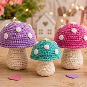Puede incluir: Tres adornos de setas de ganchillo con sombreros de color morado, turquesa y rosa, decorados con pequeños puntos blancos circulares. Los tallos están hechos de hilo color crema con un patrón de punto texturizado y se mantienen erguidos.