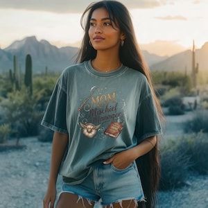 May include: A sage green short-sleeve t-shirt featuring a graphic print with the text "MOM Mischief Manager" in a stylized font. The design includes illustrations of a wand, a winged golden snitch, and a small book labeled "MOM'S MANUAL OF MISCHIEF."