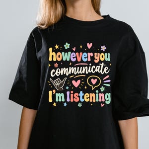 Puede incluir: Una camiseta negra con el texto colorido "however you communicate I'm listening" en una fuente divertida. El diseño incluye pequeños elementos gráficos como corazones, estrellas, flores, una mano haciendo el signo shaka y un bocadillo.