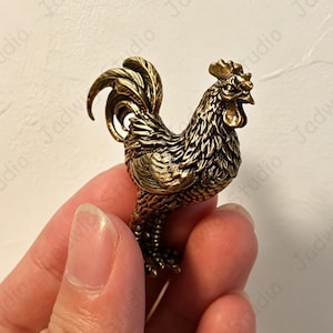 Puede incluir: Una pequeña figura tridimensional de latón que representa un gallo con plumas detalladas, una cresta prominente y una cola curvada. El metal tiene un acabado dorado antiguo bruñido con áreas oscuras que resaltan la textura del ave.