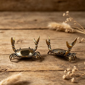 May include: Two small, metallic, brass-coloured crab figurines with raised claws. Each crab features a detailed shell texture and articulated legs, positioned to stand upright on a flat surface. The finish has a slightly aged, burnished appearance.