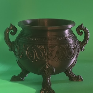May include: A black cauldron with a wide circular opening, featuring two ornate side handles and three clawed feet. The exterior surface is decorated with raised, swirling patterns and intricate detailing around the rim and base.