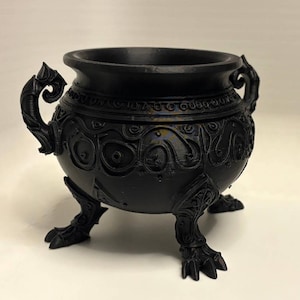 May include: A matte black cauldron featuring intricate raised scrollwork and circular patterns around the exterior. The vessel is supported by three clawed feet and includes two ornate, curved handles on opposite sides of the rim.