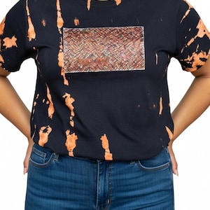 Könnte beinhalten: Ein schwarzes Kurzarm-T-Shirt mit einem Bleichmittel-Muster aus orangefarbenen vertikalen Streifen. In der Mitte der Brust befindet sich ein rechteckiger Grafikaufnäher mit einer strukturierten, mehrfarbigen Weboptik in Rot-, Braun- und Beigetönen.