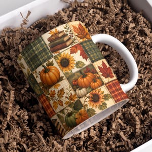 Peut inclure: Une tasse en céramique blanche ornée d'un motif patchwork d'illustrations sur le thème de l'automne, incluant des citrouilles orange, des tournesols jaunes, des feuilles d'érable rouges, des carreaux verts et une pile de livres.