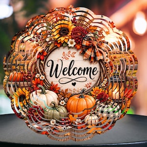 Peut inclure: Un carillon à vent circulaire en métal avec un motif automnal et le mot Welcome écrit en lettres noires. L'illustration montre des citrouilles orange et blanches, des tournesols, des feuilles d'automne, un nœud écossais et une lanterne.
