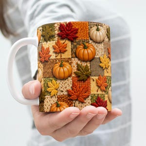 May include: A white ceramic mug featuring a patchwork design of various autumn-themed squares. The pattern includes illustrations of orange pumpkins and colourful fall leaves in shades of red, yellow, and brown against a textured fabric background.