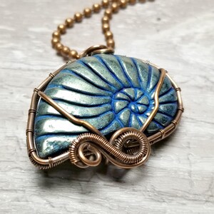 May include: A handcrafted pendant necklace featuring a blue and green nautilus shell encased in copper wire. The pendant hangs from a gold-colored beaded chain. The shell's spiral design is highlighted with blue accents, creating a unique, artistic piece.