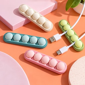 May include: Four rectangular silicone cable organizers in pastel pink, light blue, cream, and light green. Each organizer features five rounded, raised buttons designed to hold charging cables in place, with one green unit holding two white USB cables.