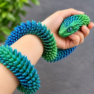 May include: A flexible articulated snake toy featuring a gradient color pattern that transitions from vibrant green at the head to a deep blue along the body. The surface is covered in numerous small, pointed, scale-like spikes throughout.