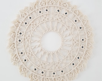 May include: A white macrame wall hanging with a circular design and black beads. The hanging has a fringe border.