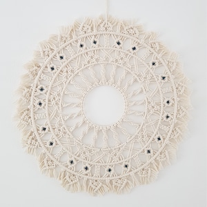 May include: A white macrame wall hanging with a circular design and black beads. The hanging has a fringe border.