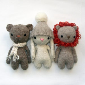 Op de afbeelding: Drie gebreide knuffels: een bruine beer met een witte sjaal, een wit konijn met een bruine hoed met een witte pompon en een bruine leeuw met een rode manen.