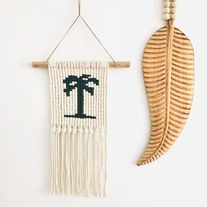 May include: A white macrame wall hanging with a black palm tree design and wooden dowel rod. A wooden leaf wall hanging with a beaded string.