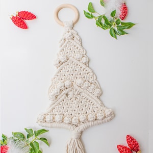 May include: A white macrame Christmas tree wall hanging with a wooden ring at the top. The tree is decorated with white pom-poms and has a long tassel at the bottom.