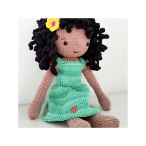 Könnte beinhalten: Handgestrickte Puppe mit dunklem Lockenhaar und einer gelben Blume. Die Puppe trägt ein hellgrünes Kleid mit rosa Akzenten und kleinen floralen Details. Die Arme und Beine der Puppe sind hellbraun.
