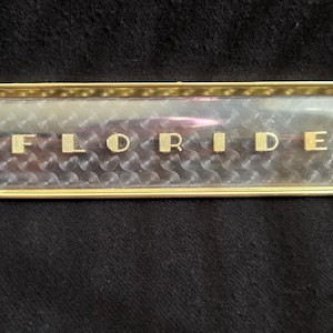 May include: A rectangular gold-toned metal emblem featuring the word FLORIDE in raised, textured gold lettering. The background behind the letters consists of a clear, reflective panel with a repeating circular, engine-turned pattern.