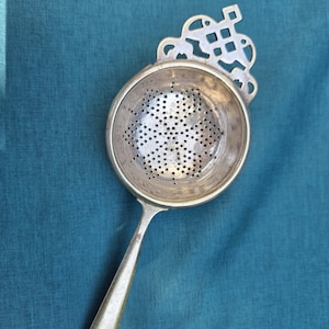 May include: A silver-toned metal tea strainer featuring a circular bowl with a perforated base and a decorative, openwork handle attachment with a geometric knot-like design. The strainer has a smooth, elongated handle extending from the bowl.