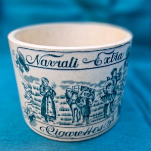Może przedstawiać: Cylindryczny ceramiczny pojemnik o kremowej podstawie z niebieskim nadrukiem. Tekst brzmi Navrati Extra Cigarettes. Ilustracja przedstawia stojącą osobę, osobę prowadzącą zwierzę juczne oraz inną osobę niosącą duży pakunek.