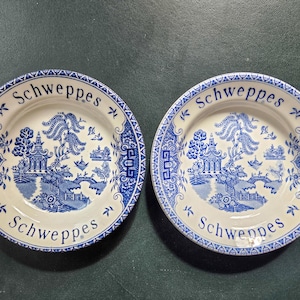 May include: Two round ceramic plates featuring a blue and white willow pattern with a pagoda, bridge, and trees. The word Schweppes is printed in blue text at the top and bottom of the rim of each plate, which also features a decorative blue border.