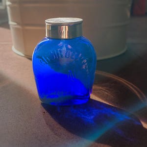 May include: A small, cobalt blue glass bottle with a rounded body and a metallic silver screw-top lid. The glass features embossed text that reads "CHOIR DE POU" in a curved pattern across the front surface, catching light to create a blue shadow.