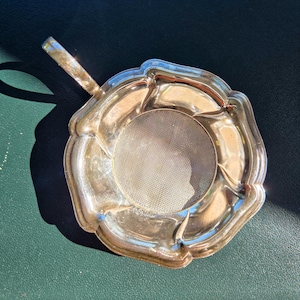 May include: A silver-toned metal tea strainer with a circular mesh centre and a scalloped, decorative rim. The strainer features a single handle extending from one side, with a polished, reflective surface that catches the light.