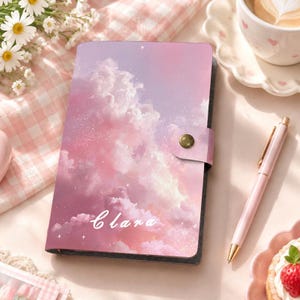 May include: A rectangular notebook with a pink and purple cloud-patterned cover featuring the name Clara in white script. The notebook has a side flap with a gold-toned snap button closure and a subtle glittery effect across the sky design.