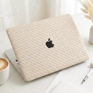 Puede incluir: Un ordenador portátil con una funda protectora de color beige con textura trenzada y un logotipo de Apple negro visible en el centro. El dispositivo está colocado sobre una superficie blanca junto a una taza de cerámica blanca con café, un bolígrafo y un cuaderno.