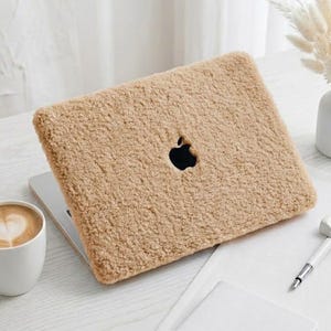 Peut inclure: Une housse d'ordinateur portable en fausse fourrure beige et duveteuse recouvrant un ordinateur fermé avec une découpe visible pour le logo Apple noir. La housse présente une matière épaisse, texturée et polaire sur le dessus.