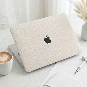 Peut inclure: Un ordinateur portable fermé avec une peau en tissu texturé de couleur crème arborant un logo Apple noir centré. L'appareil est posé sur une surface blanche à côté d'une tasse en céramique blanche contenant un café avec un motif de cœur en mousse.