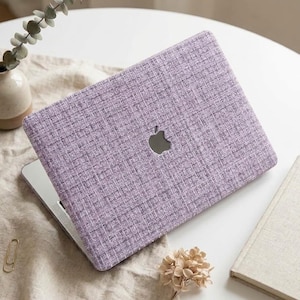 Op de afbeelding: Een laptop met een gestructureerde, lichtpaarse, stofachtige hoes met een zichtbaar weefpatroon. Het apparaat heeft een gecentreerd zilveren Apple-logo op het deksel. De laptop is gesloten en in een hoek op een neutraal gekleurd oppervlak geplaatst.