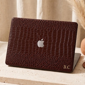 May include: A dark brown laptop case with a textured crocodile skin pattern and a cutout for the Apple logo. The bottom right corner features the gold-colored initials B.C. printed in a serif font on the surface of the protective cover.