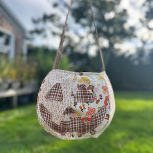 May include: A fabric pumpkin-shaped treat bag featuring a cream-colored base with a repeating pattern of cartoon children and flowers. The jack-o'-lantern face is appliqued with brown and white plaid fabric, and it includes a thin brown fabric handle.