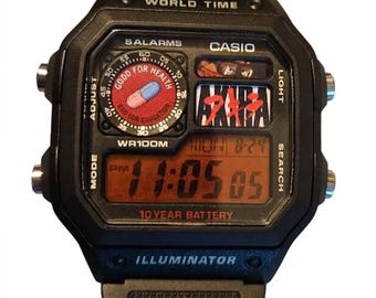 Puede incluir: Un reloj de pulsera digital negro con caja cuadrada y correa de resina. La esfera muestra el texto 'WORLD TIME', 'CASIO', '5 ALARMS', 'WR100M', '10 YEAR BATTERY' e 'ILLUMINATOR'. Incluye un gráfico de una pastilla y el texto 'AKIRA'.