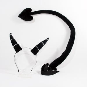 May include: A black headband featuring two plush horns with white horizontal stripes, paired with a long, curved black plush tail that ends in a heart-shaped tip. The tail includes a black fabric strap at the base for attachment purposes.