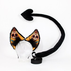May include: A set consisting of a headband with two plush cat ears and a long black tail. The ears feature a yellow fabric with a printed sugar skull design in red, black, and white, accented with black faux fur. The tail has a heart-shaped tip.