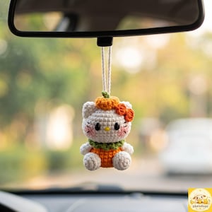May include: A white crocheted cat figure hanging from a white cord, featuring a small orange pumpkin on its head, an orange bow, a green collar, and an orange pumpkin-shaped body. The cat has black eyes, a yellow nose, and pink cheeks.
