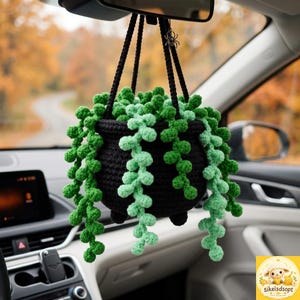 May include: A crocheted black cauldron hanging from a rearview mirror, filled with trailing light and dark green beaded vines. The plant features small, round, textured leaves cascading over the sides, and a small silver spider charm is attached.