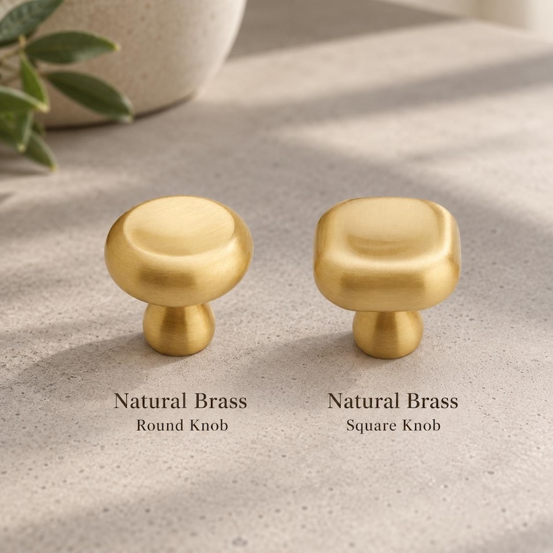 Może przedstawiać: Dwie gałki z naturalnego mosiądzu o szczotkowanym wykończeniu. Jedna ma okrągłą g&oacute;rę, oznaczoną jako Natural Brass Round Knob. Druga ma kwadratową g&oacute;rę z zaokrąglonymi krawędziami, oznaczoną jako Natural Brass Square Knob. Obie mają podstawę.