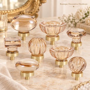 May include: A collection of eight cabinet knobs featuring light amber-colored glass tops in various shapes, including round, square, and fluted designs, each mounted on a circular brushed brass base. The text Boutique Decorative Hardware is visible.