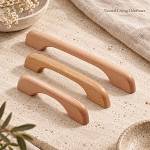 Puede incluir: Tres tiradores de armario de madera de color claro y diferentes longitudes dispuestos en diagonal. Cada tirador presenta un diseño liso y curvado con extremos redondeados. El texto Natural Living Hardware es visible en la esquina superior derecha.