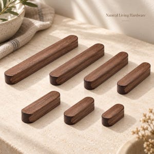 Puede incluir: Una colección de siete tiradores de armario rectangulares de madera marrón oscuro de varias longitudes, dispuestos en dos filas. Cada pieza presenta un acabado de grano de madera maciza lisa con bordes redondeados. El texto Natural Living Hardware es visible.