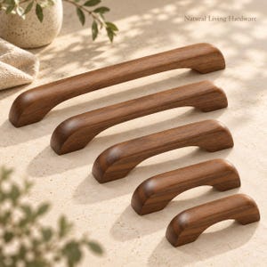 May include: Five dark brown wooden cabinet pulls of varying lengths arranged in a diagonal row. Each handle features a smooth, curved design with rounded ends. The text Natural Living Hardware is visible in the upper right corner of the image.