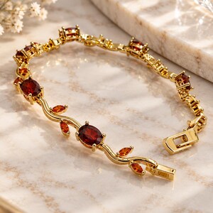 Puede incluir: Una pulsera de tono dorado con un diseño de enredadera, adornada con piedras granate ovaladas de color rojo oscuro que alternan con gemas naranjas más pequeñas en talla marquesa. Los eslabones metálicos imitan tallos y hojas, con un cierre de caja rectangular.