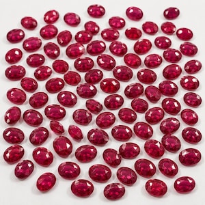 Puede incluir: Una gran colección de piedras preciosas rojas sueltas, talladas en óvalo con una superficie facetada. Cada piedra muestra un tono carmesí profundo y vibrante con un acabado pulido, esparcidas sobre una superficie blanca lisa.