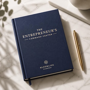 May include: A navy blue hardback planner with gold foil text that reads THE ENTREPRENEUR'S COMMAND CENTER and BLOOMLINE PLANNERS with a floral logo. A gold metal pen rests beside the planner, which features a matching navy blue ribbon bookmark.