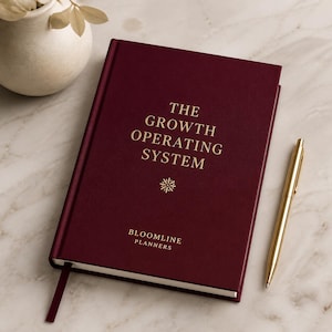 May include: A deep burgundy hardcover planner titled THE GROWTH OPERATING SYSTEM in gold foil lettering, featuring a small starburst icon and the text BLOOMLINE PLANNERS at the bottom, accompanied by a gold metal pen with a matching ribbon bookmark.